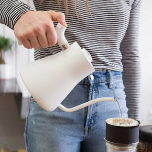 Kéz, amely fehér, keskeny csőrű vízforralóból vizet önt egy kávészűrőbe, az EspressoGear része, minden a kávéról.