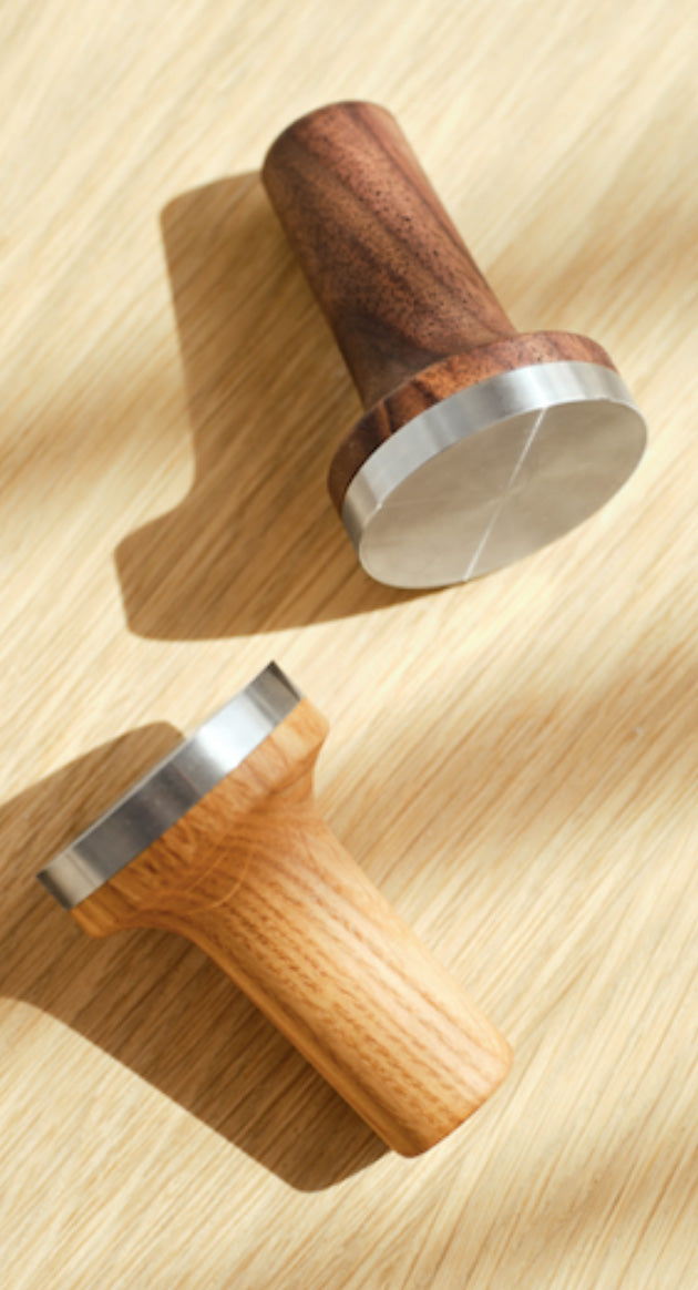 Två kaffetampers med trähandtag och rostfria stålytor på ljus träyta i naturligt ljus.